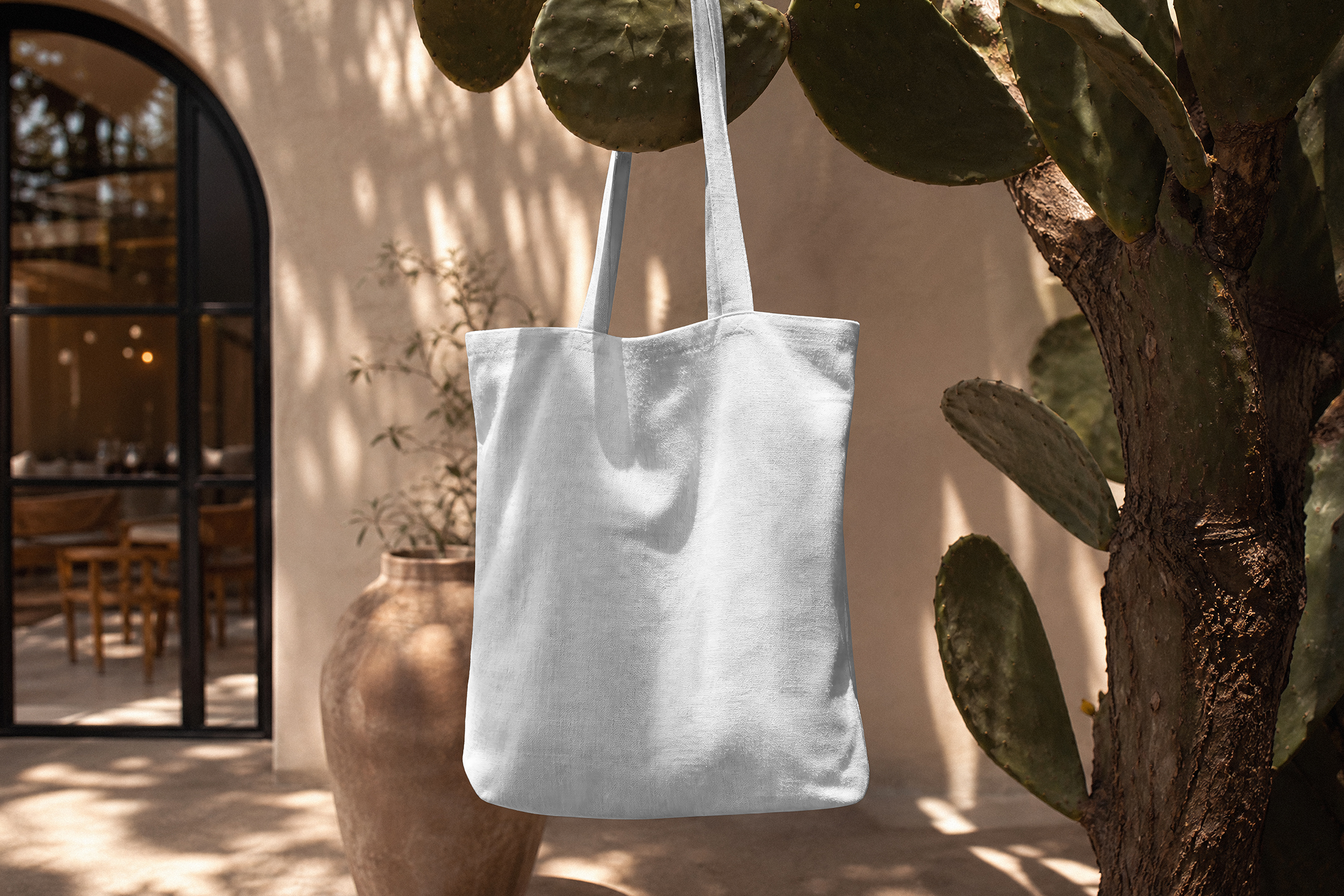 A blank white canvas tote bag hanging from a prickly pear cactus branch in front of a warm-toned Mediterranean building.