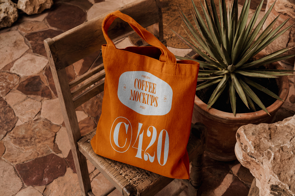 An orange canvas tote bag with custom white typography resting on a rustic wooden chair next to a potted agave plant on a stone floor.