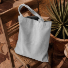 A plain light grey canvas tote bag sitting on a rustic wooden woven chair next to a potted pointed plant on a stone tile floor.