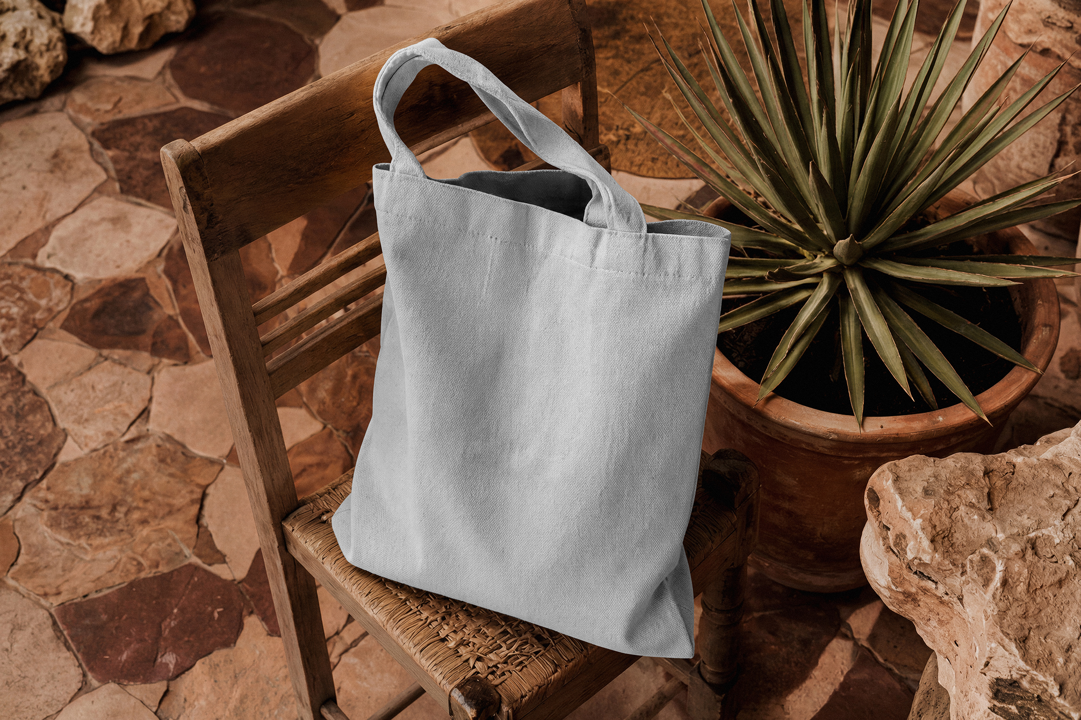 A plain light grey canvas tote bag sitting on a rustic wooden woven chair next to a potted pointed plant on a stone tile floor.