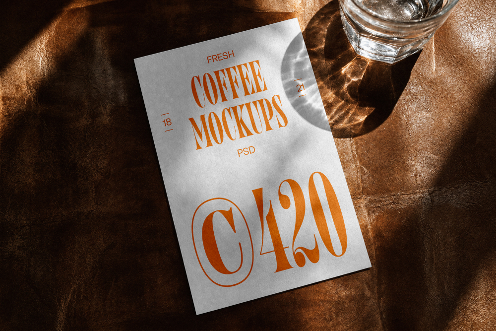 A white vertical stationery card with bold orange coffee branding typography, placed on a dark brown leather surface beside a glass of water in direct sunlight.