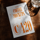 A white vertical stationery card with bold orange coffee branding typography, placed on a dark brown leather surface beside a glass of water in direct sunlight.