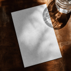 Top-down view of a blank white rectangular card lying on a dark brown textured leather surface beside a clear glass of water under bright sunlight.