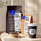 A set of coffee packaging mockups on a stone counter, featuring a dark brown coffee bag, an orange bag, a patterned paper cup, and a small bowl of roasted coffee beans.
