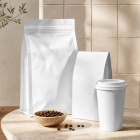 Blank white coffee packaging mockups consisting of two stand-up pouches and a takeaway paper cup next to a small bowl of coffee beans on a stone table.