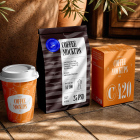 A set of coffee packaging mockups, including an orange paper cup, a dark brown coffee pouch, and an orange box, arranged on a tiled floor in sunlight.