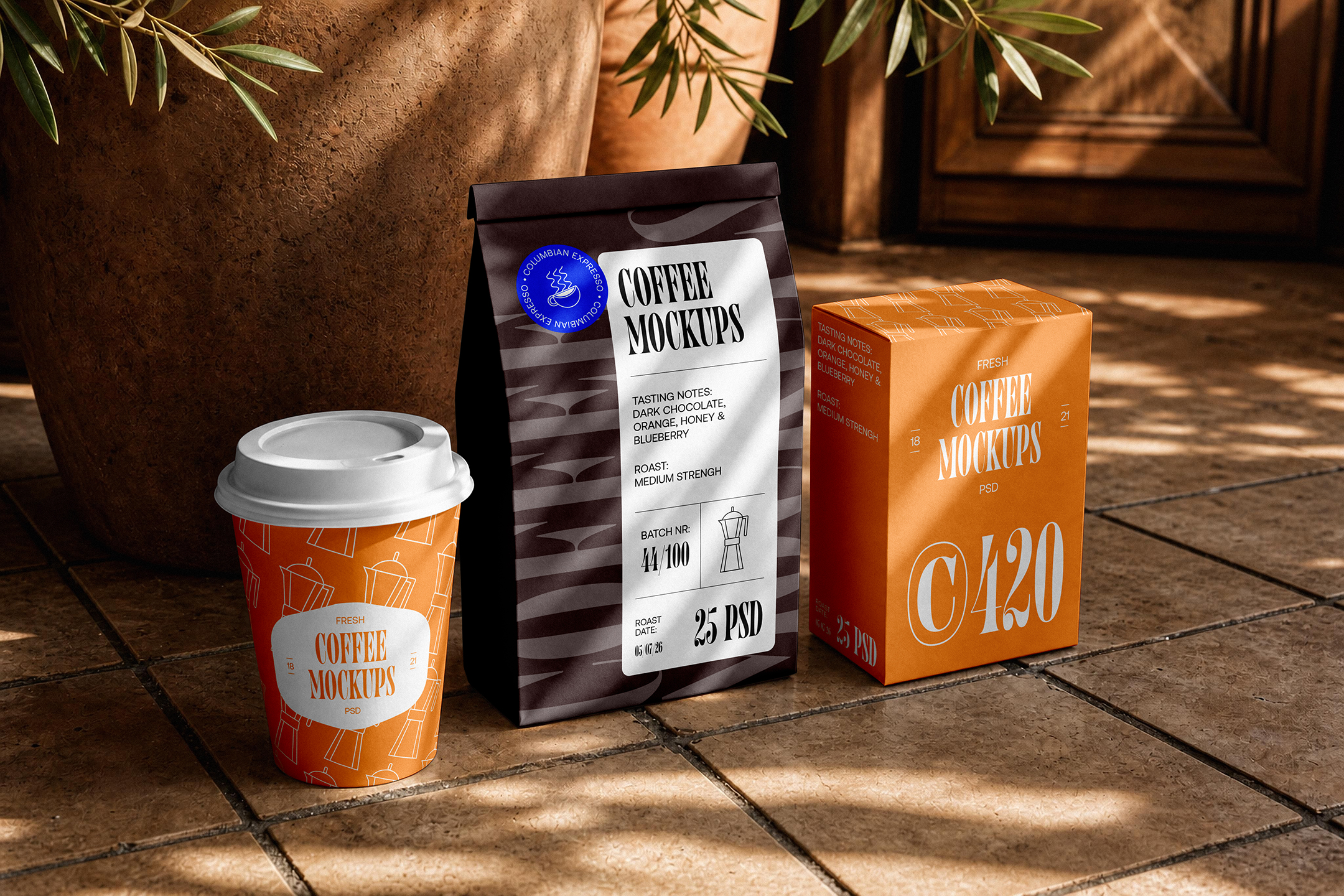 Coffee-Branding-Mockups-03-CMV2-A A set of coffee packaging mockups, including an orange paper cup, a dark brown coffee pouch, and an orange box, arranged on a tiled floor in sunlight.