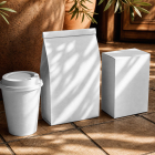 A blank white paper coffee cup, folded paper bag, and rectangular box resting on a tiled floor in warm dappled sunlight next to a terracotta planter.