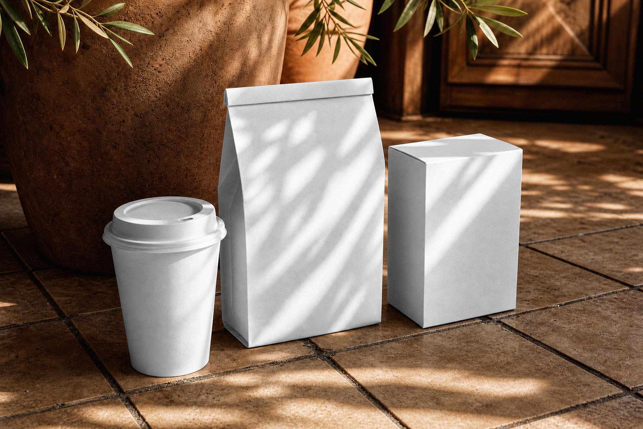 A blank white paper coffee cup, folded paper bag, and rectangular box resting on a tiled floor in warm dappled sunlight next to a terracotta planter.