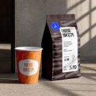 An orange takeaway paper coffee cup standing next to a dark brown coffee bag on a concrete floor with sunlight casting window shadows.