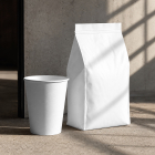A blank white paper coffee cup without a lid standing next to a tall white coffee bag on a concrete floor with geometric sunlight shadows.
