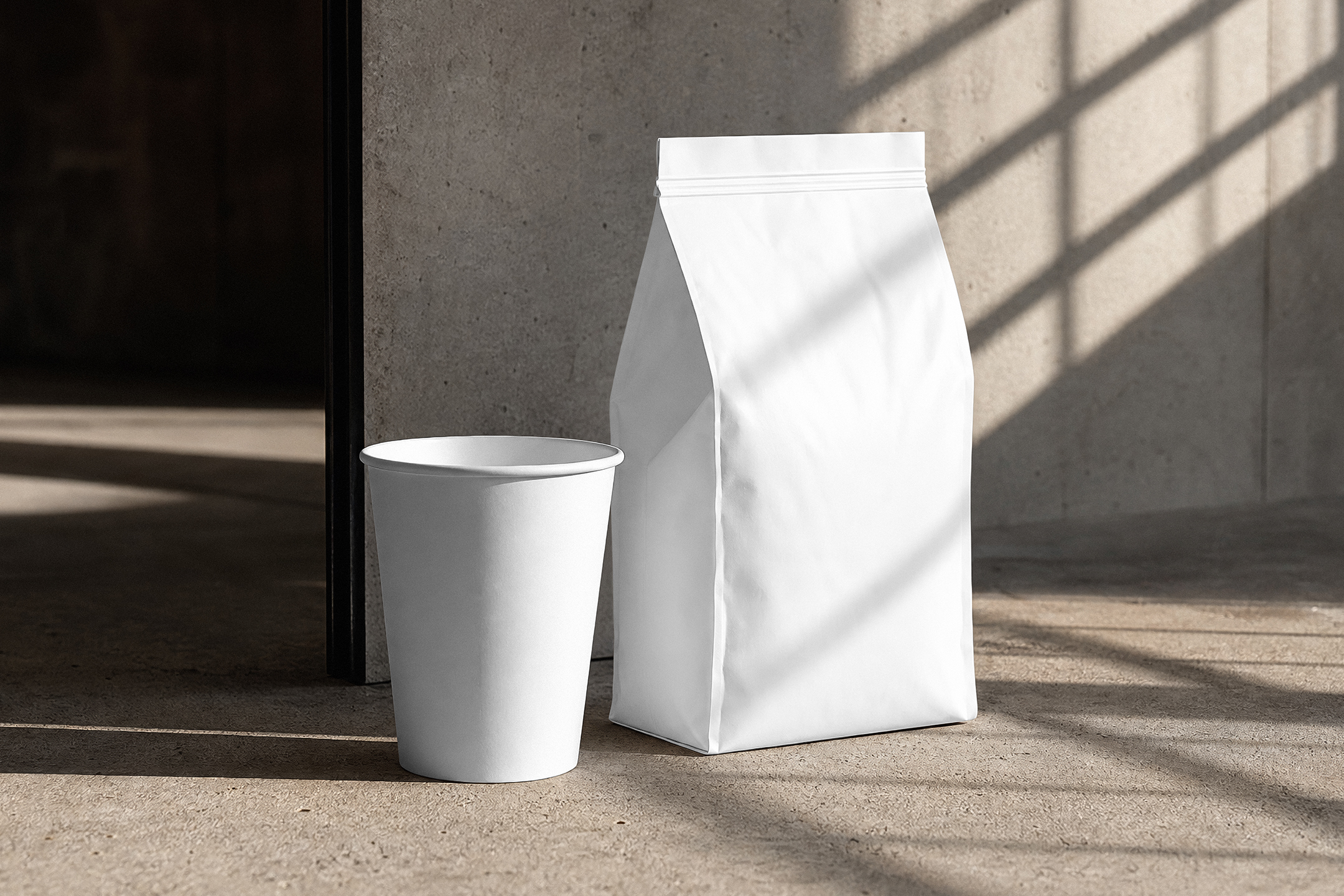 A blank white paper coffee cup without a lid standing next to a tall white coffee bag on a concrete floor with geometric sunlight shadows.