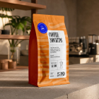 An orange stand-up coffee pouch with a custom label placed on a stone café countertop, with coffee machines blurred in the background.