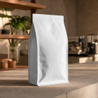 A blank white stand-up coffee pouch standing on a stone countertop in a cozy, sunlit café setting with wooden shelves in the background.