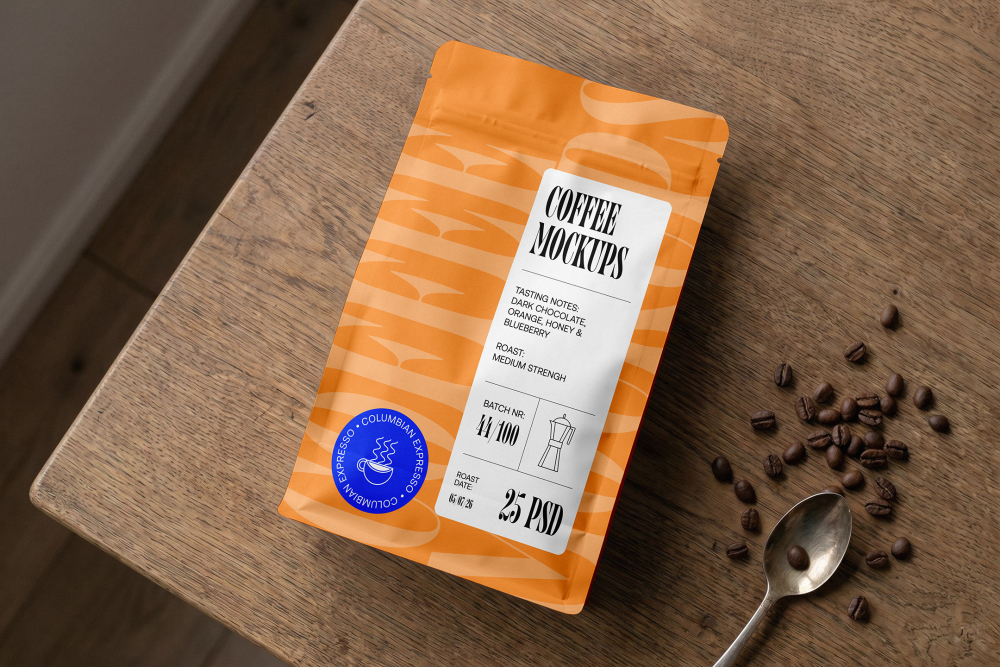 Top-down view of an orange coffee bag lying flat on a wooden table, surrounded by scattered coffee beans and a metal spoon.