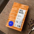 Top-down view of an orange coffee bag lying flat on a wooden table, surrounded by scattered coffee beans and a metal spoon.