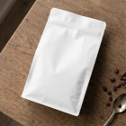 Flat lay view of a blank white zip-lock coffee pouch lying on a wooden table beside scattered roasted coffee beans and a vintage spoon.