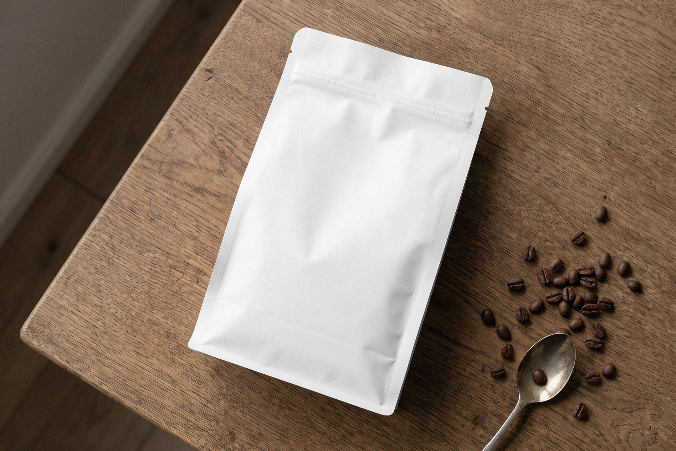 Flat lay view of a blank white zip-lock coffee pouch lying on a wooden table beside scattered roasted coffee beans and a vintage spoon.