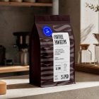 A dark brown coffee bean bag standing on a concrete kitchen counter alongside coffee brewing gear, including a pour-over dripper and espresso machine.