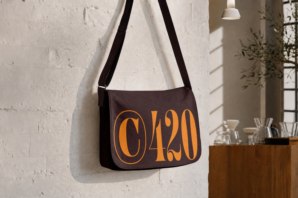 A dark brown canvas messenger bag printed with bold orange "C420" typography hanging on a white brick wall.