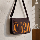 A dark brown canvas messenger bag printed with bold orange "C420" typography hanging on a white brick wall.