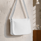 A blank white canvas crossbody messenger bag hanging on a textured white brick wall in a modern café setting.