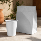 A blank white takeaway paper cup and folded paper bag sitting on a concrete surface next to a terracotta plant pot in dappled sunlight.