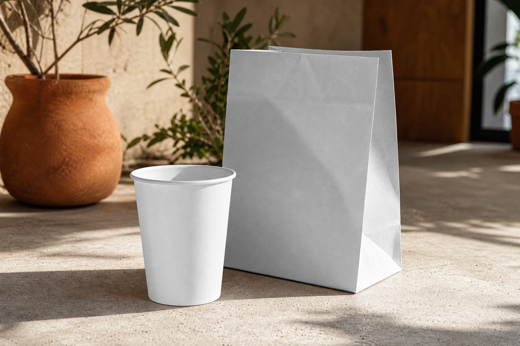 A blank white takeaway paper cup and folded paper bag sitting on a concrete surface next to a terracotta plant pot in dappled sunlight.