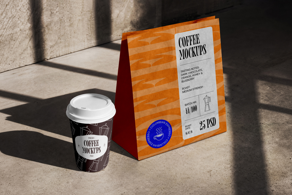A dark paper coffee cup with a white lid and an orange paper shopping bag resting on a concrete surface under harsh sunlight.