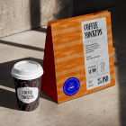 A dark paper coffee cup with a white lid and an orange paper shopping bag resting on a concrete surface under harsh sunlight.