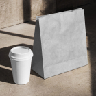 High-angle view of a blank white paper coffee cup with a lid standing next to a folded white paper bag on a concrete floor with strong geometric shadows.