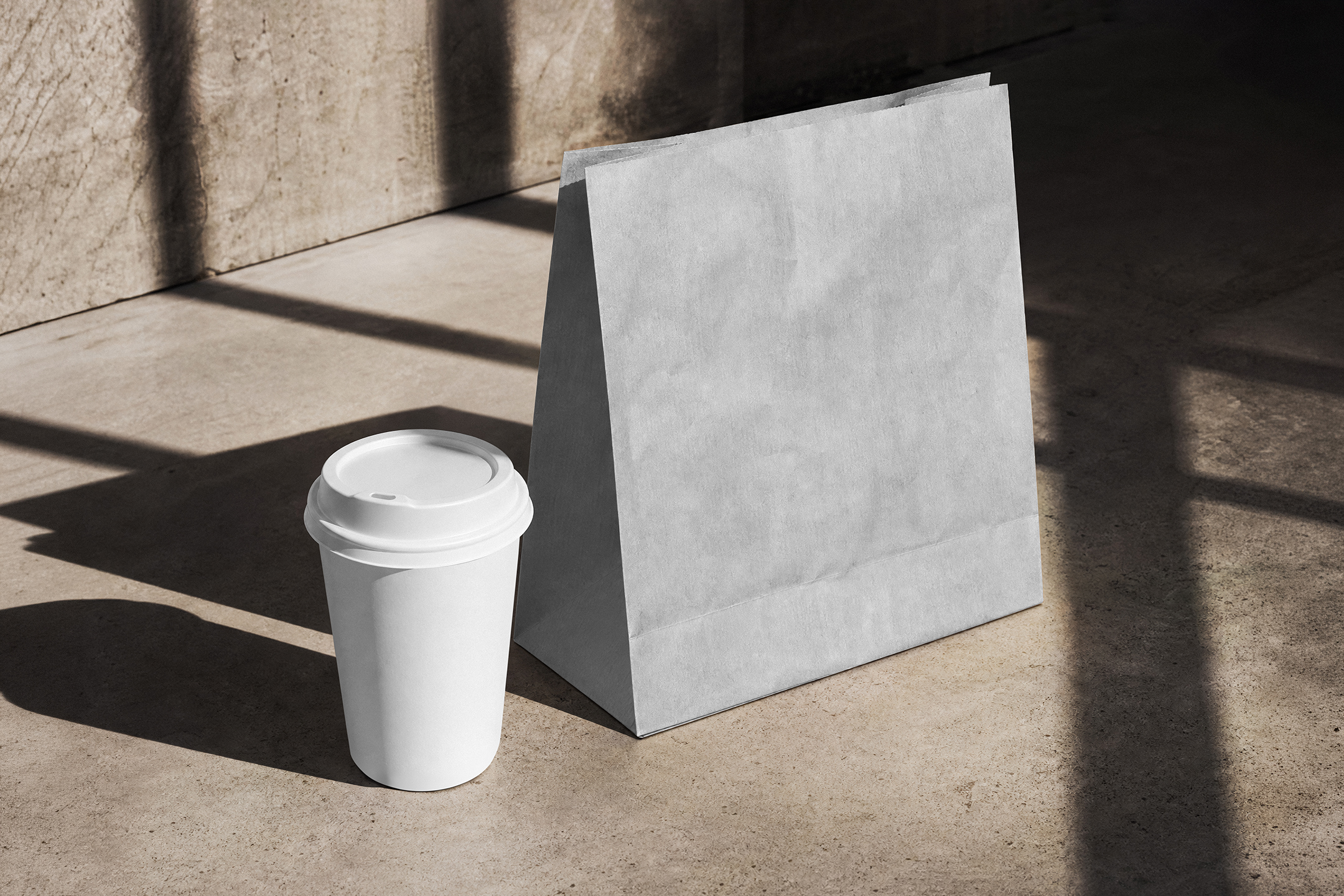 High-angle view of a blank white paper coffee cup with a lid standing next to a folded white paper bag on a concrete floor with strong geometric shadows.
