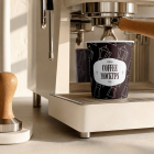 A dark paper coffee cup sitting directly under the group head of a modern cream-colored espresso machine, next to a wooden tamper.