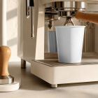 A blank white paper cup placed under the portafilter of a modern beige espresso machine beside a wooden tamper.