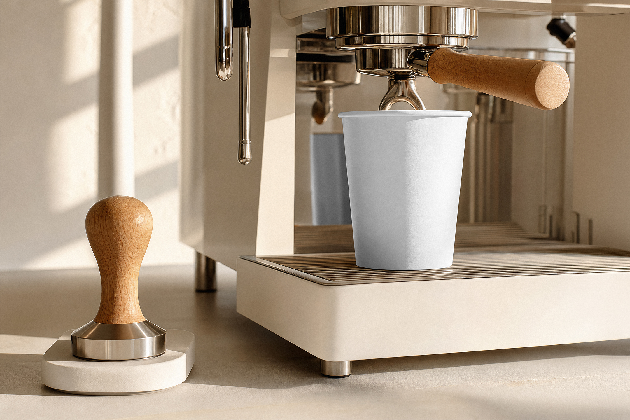 A blank white paper cup placed under the portafilter of a modern beige espresso machine beside a wooden tamper.