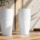 Two tall stacks of blank white paper coffee cups standing side-by-side on a wooden counter in a bright, sunlit room.