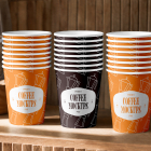 Three stacks of paper coffee cups—two orange and one dark brown in the center—lined up on a wooden shelf in front of a wooden slatted wall.
