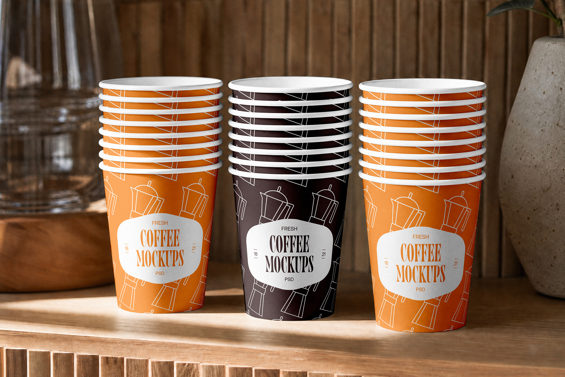 Paper-Coffee-Cup-Mockup-03-CMV2-A Three stacks of paper coffee cups—two orange and one dark brown in the center—lined up on a wooden shelf in front of a wooden slatted wall.