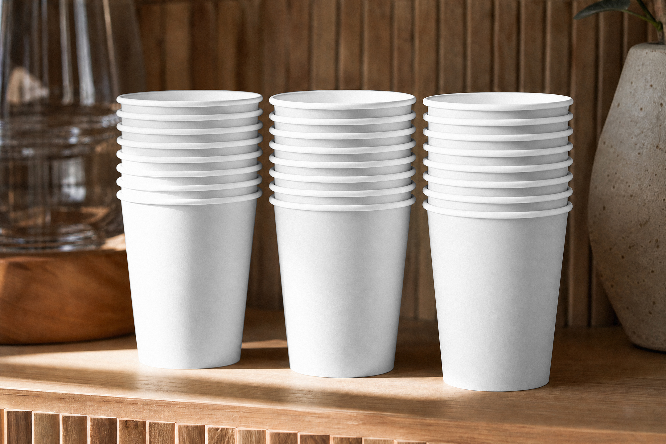 Three stacks of blank white paper coffee cups lined up on a wooden shelf against a slatted wooden wall.