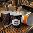 An espresso machine pouring coffee into a dark paper cup on a stainless steel drip tray, with two additional branded paper cups beside it.