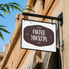 A hanging white outdoor store sign mounted on a textured stone building façade under a clear blue sky, displaying a dark brown logo badge.