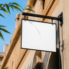 A blank white rectangular hanging store sign with a black metal frame mounted on the rustic stone wall of a building under a clear blue sky.
