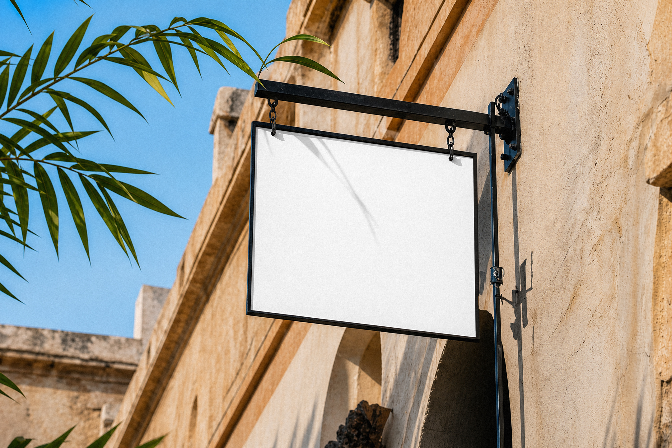 A blank white rectangular hanging store sign with a black metal frame mounted on the rustic stone wall of a building under a clear blue sky.