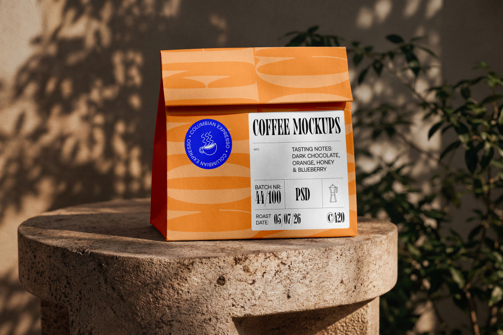 An orange paper takeaway coffee bag with a detailed product label and blue sticker, standing on a round stone pedestal outdoors in dappled sunlight.