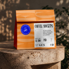 An orange paper takeaway coffee bag with a detailed product label and blue sticker, standing on a round stone pedestal outdoors in dappled sunlight.