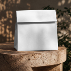 A blank white folded takeaway paper bag sitting on a carved stone stool against a warm wall with dappled plant shadows.