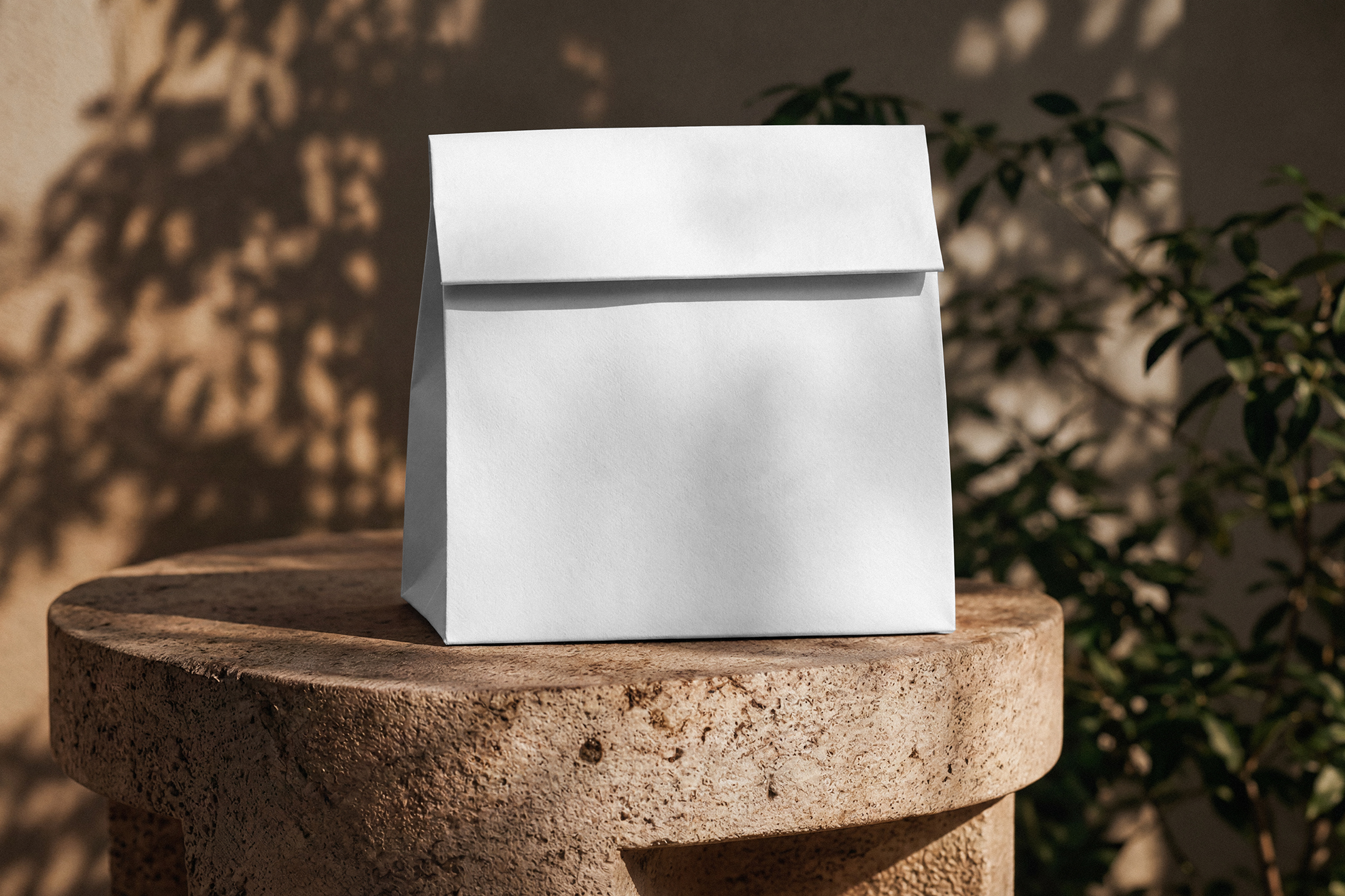 A blank white folded takeaway paper bag sitting on a carved stone stool against a warm wall with dappled plant shadows.