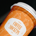 Close-up angled view of an orange paper coffee cup with a white lid, featuring a Moka pot pattern and a white badge reading "Fresh Coffee Mockups".