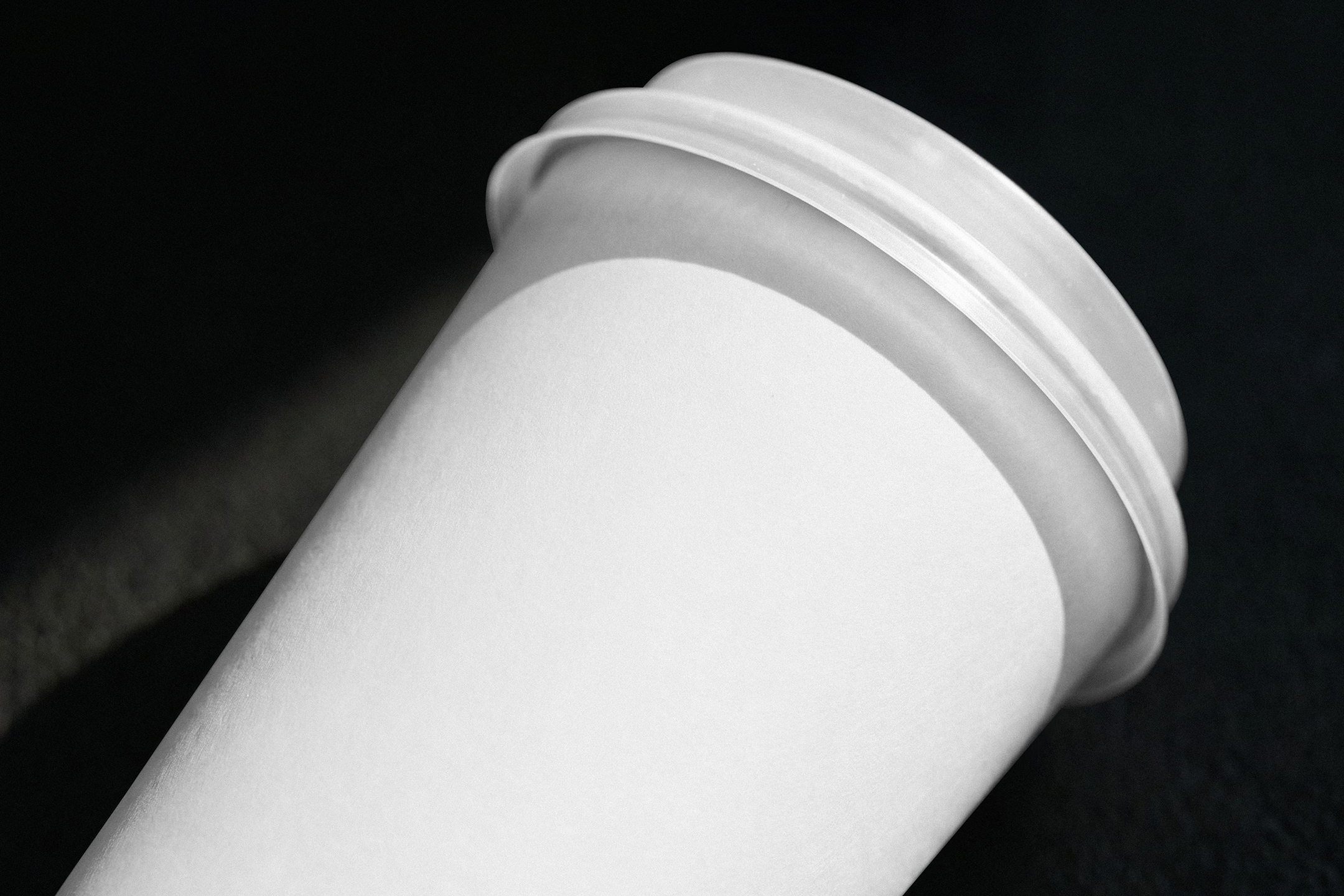 Close-up view of a blank white paper coffee cup with a lid resting at an angle on a dark, textured surface.
