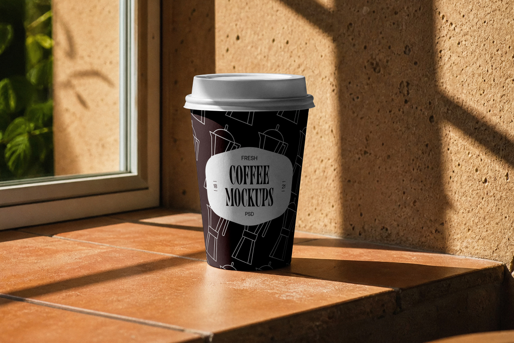 A dark brown paper coffee cup with a white lid resting on a terracotta tiled windowsill under warm sunlight and foliage shadows.